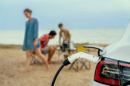 Foto de Viaje de vacaciones familiar alternativo que viaja por la playa con batería de recarga de coche eléctrico desde la estación de carga de EV con la familia borrosa disfrutando del fondo del paisaje marino. Perpetuo - Imagen libre de derechos