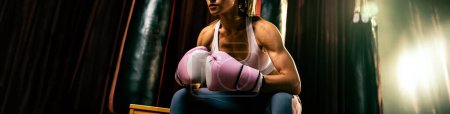 Téléchargez les photos : Femme asiatique Muay boxeuse thaïlandaise ou kickboxing prendre une courte pause assis avec ses gants sur la salle de gym avec du matériel de boxe en arrière-plan. Forte et musclée sportive du corps. Éperon - en image libre de droit