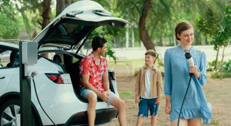 Foto de Vacaciones de viaje en familia con vehículo eléctrico, encantadora familia recarga coche EV con energía verde y limpia. Viajes en coche naturales y ecológicos para un medio ambiente sostenible. Perpetuo - Imagen libre de derechos