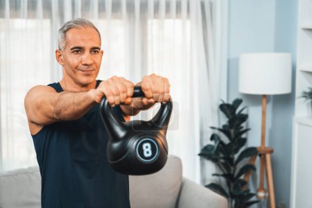 Photo for Athletic and sporty senior man engaging in body workout routine with lifting kettlebell at home as concept of healthy fit body with body weight lifestyle after retirement. Clout - Royalty Free Image