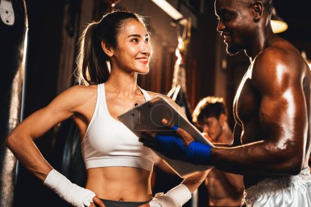 Téléchargez les photos : Femme asiatique Muay boxeuse thaïlandaise et son entraîneur de boxe personnelle discuter de ses progrès physiques dans la salle de gym reflétant son engagement à la croissance musculaire de son corps et la performance de boxe. Impulsion - en image libre de droit