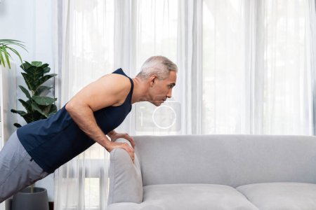Foto de Hombre mayor atlético y activo que utiliza muebles para dirigir el músculo eficaz con empuje hacia arriba en el ejercicio en el hogar como concepto de estilo de vida corporal en forma saludable después de la jubilación. Clout. - Imagen libre de derechos