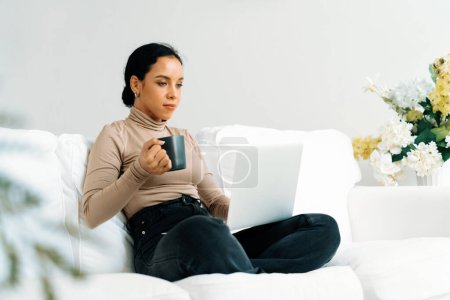Foto de Mujer feliz bebiendo café en un sofá en casa para el descanso y la relajación crucial. Retrato de una joven afroamericana sosteniendo una taza. - Imagen libre de derechos