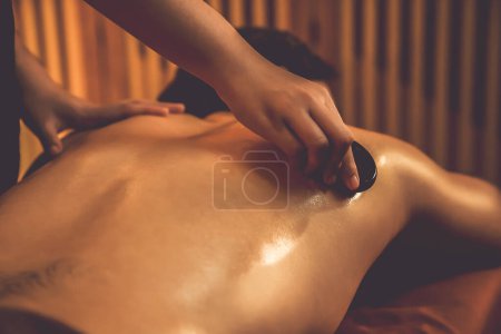 Téléchargez les photos : Massage aux pierres chaudes au salon de spa dans un complexe de luxe avec lumière chaude aux bougies, client homme heureux profitant d'un massage aux pierres de basalte spa glisse sur le corps avec une chaleur apaisante. Tranquillité - en image libre de droit