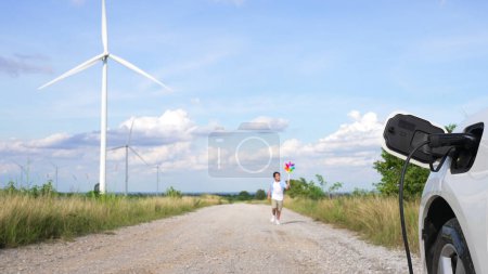 Téléchargez les photos : Progressif jeune garçon jouant jouet moulin à vent à côté de voiture EV au parc d'éoliennes. Générateur électrique à partir du vent par éolienne sur le côté campagne. - en image libre de droit