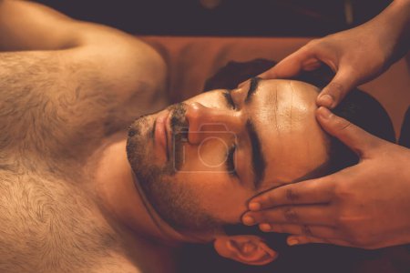 Téléchargez les photos : Homme caucasien profiter relaxant anti-stress massage de la tête et choyer beauté faciale loisirs de la peau dans la bougie chaude éclairage spa salon ambiant dans un complexe de luxe ou un hôtel. Tranquillité - en image libre de droit