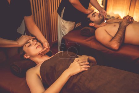 Téléchargez les photos : Couple client appréciant relaxant anti-stress massage de la tête et choyer beauté faciale loisirs de la peau dans la bougie chaude éclairage spa salon d'ambiance dans un complexe de luxe ou un hôtel. Tranquillité - en image libre de droit