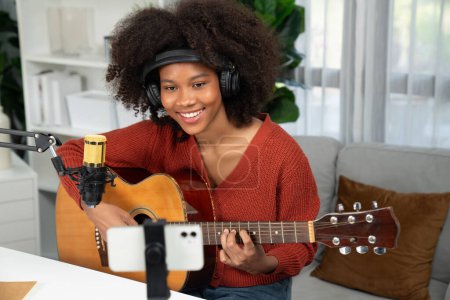 Foto de Anfitrión de canal en el músico de jóvenes afroamericanos tocando la guitarra junto con el canto, la radiodifusión en el teléfono inteligente en el estudio. Decoración de los auriculares del equipo y micrófono de grabación. Tastemaker. - Imagen libre de derechos