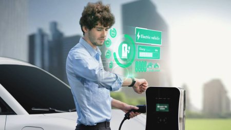 Téléchargez les photos : Homme d'affaires vérifier l'état de la batterie de voiture EV sur la recharge hologramme smartwatch de la station de charge ESG parc vert. Style de vie urbain moderne et utilisation écologique de la voiture électrique pour réduire le CO2. Pertinence - en image libre de droit