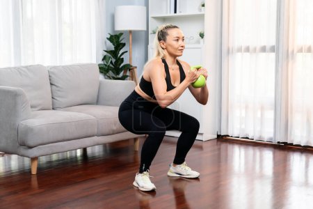 Photo for Athletic and sporty senior woman engaging in leg day training session with squat and bodyweight kettle ball at home exercise as concept of healthy fit body lifestyle after retirement. Clout - Royalty Free Image