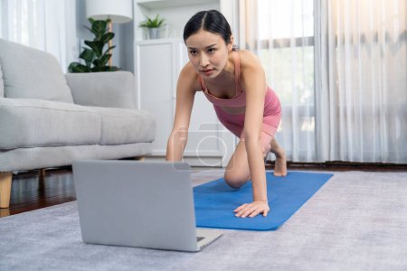 Téléchargez les photos : Femme asiatique en vêtements de sport faisant burpee sur tapis d'exercice comme routine d'entraînement à domicile. Fille attrayante s'engager dans sa poursuite d'un mode de vie sain avec la vidéo de formation d'exercice en ligne. Vigoureux - en image libre de droit