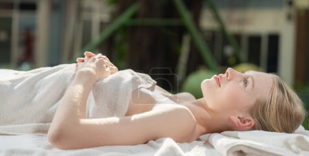 Photo for Portrait of beautiful caucasian girl in white towel lies on spa bed peacefully at outdoor surrounded by relaxing and calming environment while waiting for body massage. Blurred background. Tranquility - Royalty Free Image