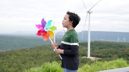 Téléchargez les photos : Petit garçon asiatique progressiste jouant avec un jouet d'éolienne dans le parc d'éoliennes, champ vert au-dessus de la colline. Énergie verte produite à partir d'éoliennes électriques renouvelables. - en image libre de droit