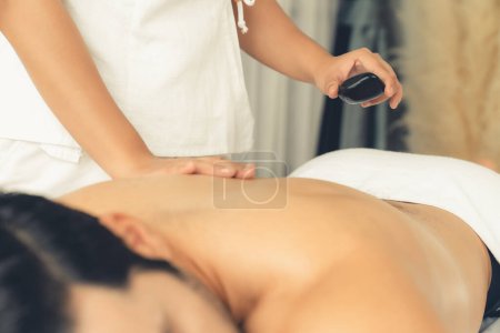 Téléchargez les photos : Massage aux pierres chaudes au salon de spa dans un complexe de luxe avec lumière du jour sérénité ambiante, homme heureux client profitant d'un massage aux pierres de basalte spa glisser sur le corps avec une chaleur apaisante. Tranquillité - en image libre de droit