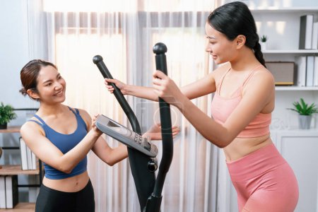Téléchargez les photos : Femme asiatique athlétique énergique et forte fonctionnant sur une machine elliptique à la maison avec un copain ou un entraîneur d'entraînement. Poursuite d'ajustement physique et l'engagement à un mode de vie sain. Vigoureux - en image libre de droit