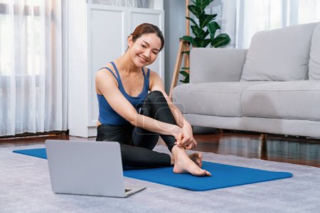 Foto de Mujer asiática en ropa deportiva sentada en el piso de la casa mientras ve videos de ejercicios en línea en el portátil. Chica atractiva participar en su búsqueda de un estilo de vida saludable. Vigoroso - Imagen libre de derechos