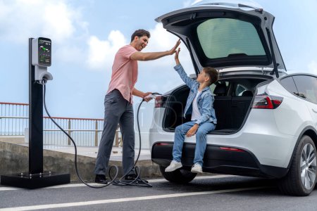 Family road trip vacation traveling by the sea with electric car, father and son high five after reach destination at EV charging station by the seashore. Eco-friendly car for environment. Perpetual
