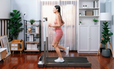 Téléchargez les photos : Pleine longueur vue latérale de femme asiatique énergique et forte course à pied machine à la maison. Poursuite de la forme physique et l'engagement à un mode de vie sain avec l'entraînement à domicile et la formation. Vigoureux - en image libre de droit