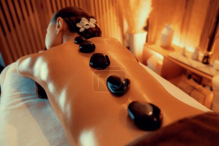 Téléchargez les photos : Massage aux pierres chaudes au salon de spa dans un complexe de luxe avec lumière chaude à la bougie, une cliente heureuse profitant d'un massage aux pierres de basalte spa glisse sur le corps avec une chaleur apaisante. Tranquillité - en image libre de droit