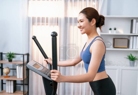 Téléchargez les photos : Femme asiatique athlétique énergique et forte qui court sur une machine elliptique à la maison. Poursuite de la forme physique et l'engagement à un mode de vie sain avec l'entraînement à domicile et la formation. Vigoureux - en image libre de droit