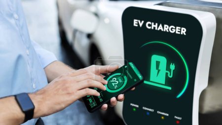 Photo for Businessman holding smartphone display battery status interface by smart EV mobile application while EV car recharging electricity from charging station in car park. Peruse - Royalty Free Image