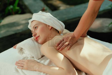 Photo for Beautiful young woman received a back massage on a spa bed from professional masseuse. Attractive female relaxes deeply by skilled hands of the massage therapist. Surrounded with nature. Tranquility. - Royalty Free Image
