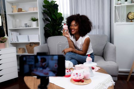 Photo for Beautiful young African blogger presenting piece of cupcake in concept special cuisine with video record on smartphone. Content creating of social media with favorite sweets bakery dish. Tastemaker. - Royalty Free Image