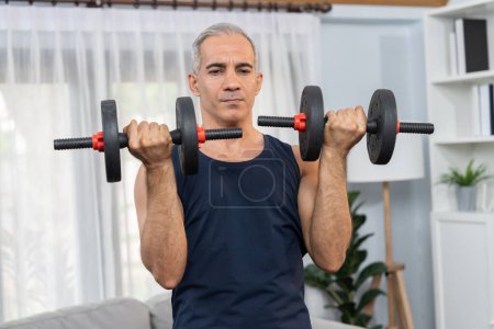 Photo for Athletic and sporty senior man engaging in body workout routine with lifting dumbbell at home as concept of healthy fit body with body weight lifestyle after retirement. Clout - Royalty Free Image