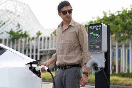 Photo for Young man put EV charger to recharge electric cars battery from charging station in city commercial parking lot. Rechargeable EV car for sustainable environmental friendly urban travel. Expedient - Royalty Free Image