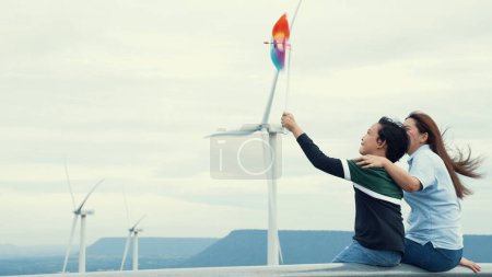 Téléchargez les photos : Progressiste mère heureuse et son fils au parc d'éoliennes. Générateur électrique à partir du vent par éolienne sur le côté campagne avec colline et montagne à l'horizon. - en image libre de droit