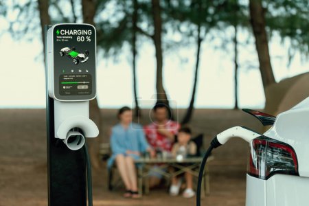 Photo for Alternative family camping trip traveling by the beach with electric car recharging battery from EV charging station with blurred family enjoying the seascape campsite background. Perpetual - Royalty Free Image