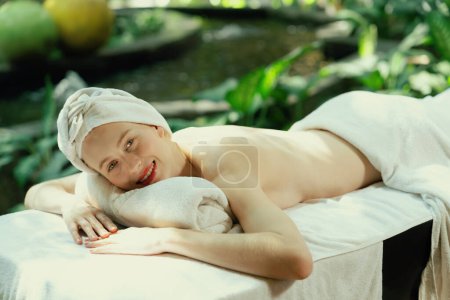 Foto de Hermosa joven mujer se relaja en una cama de spa rodeada de naturaleza. listo para un masaje corporal. Atractiva hembra en toalla blanca tumbada en paz durante la espera de masaje corporal. De cerca. Tranquilidad - Imagen libre de derechos