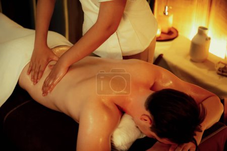 Téléchargez les photos : Homme caucasien client appréciant relaxant massage spa anti-stress et dorloter avec beauté loisirs de la peau dans la bougie chaude éclairage spa salon ambiant à la station de luxe ou hôtel. Tranquillité - en image libre de droit