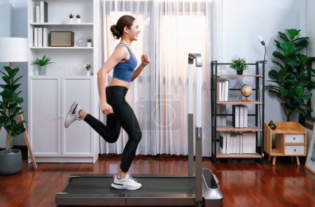 Téléchargez les photos : Pleine longueur vue latérale de femme asiatique énergique et forte course à pied machine à la maison. Poursuite de la forme physique et l'engagement à un mode de vie sain avec l'entraînement à domicile et la formation. Vigoureux - en image libre de droit