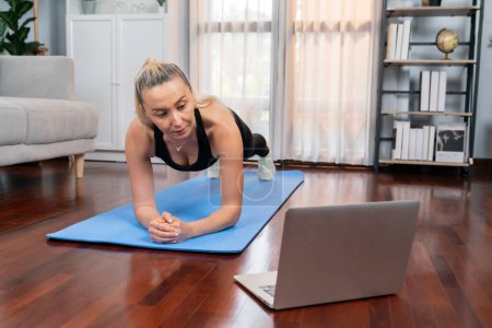 Foto de Mujer mayor atlética y deportiva planking en la alfombra de ejercicio de fitness mientras ve video de fitness en línea en el ejercicio en casa como concepto de estilo de vida corporal saludable después de la jubilación. Clout. - Imagen libre de derechos