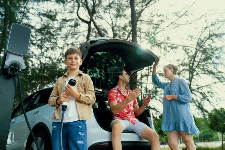 Photo for Little boy holding EV charger and point at camera with his family sitting on the trunk in background. Road trip travel with alternative energy charging station for eco-friendly car concept. Perpetual - Royalty Free Image