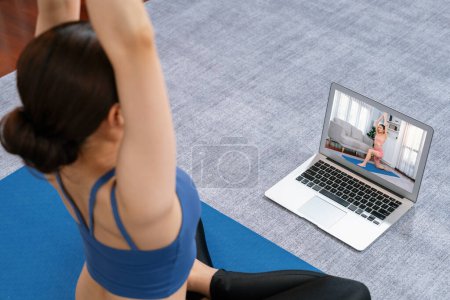 Photo for Asian woman in sportswear doing yoga exercise on fitness mat as her home workout training routine. Healthy body care lifestyle woman watching online yoga video on laptop. Vigorous - Royalty Free Image
