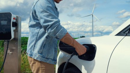 Téléchargez les photos : Homme progressiste avec sa voiture électrique, la voiture EV recharge l'énergie de la station de recharge sur champ vert avec éolienne comme concept d'énergie durable future. Véhicule électrique avec générateur d'énergie. - en image libre de droit