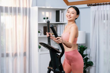 Téléchargez les photos : Femme asiatique athlétique énergique et forte qui court sur une machine elliptique à la maison. Poursuite de la forme physique et l'engagement à un mode de vie sain avec l'entraînement à domicile et la formation. Vigoureux - en image libre de droit