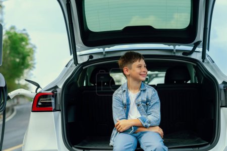 Photo for Little boy sitting on car trunk while recharging eco-friendly electric car from EV charging station. EV car road trip travel concept for alternative transportation powered sustainable energy.Perpetual - Royalty Free Image