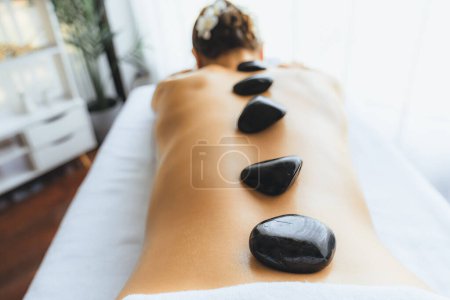 Téléchargez les photos : Massage aux pierres chaudes au salon de spa dans un complexe de luxe avec lumière du jour sérénité ambiante, femme heureuse client profitant d'un massage aux pierres de basalte spa glisser sur le corps avec une chaleur apaisante. Tranquillité - en image libre de droit
