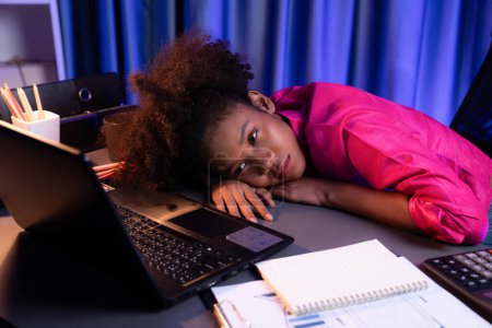 Foto de Mujer africana freelancer sintiéndose probada y tomar una siesta en el escritorio alrededor de la computadora portátil y estacionaria, esperando a continuar el trabajo del proyecto hasta que duerma en el escritorio. Concepto de vida laboral en el hogar. Tastemaker. - Imagen libre de derechos
