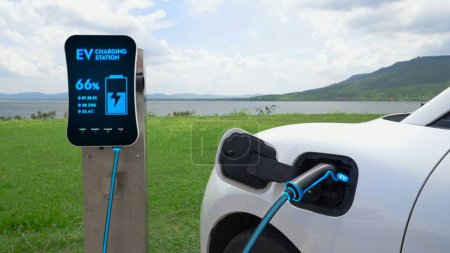 Téléchargez les photos : Voiture électrique branchée avec borne de recharge pour recharger la batterie par câble chargeur EV avec fond nature et lac. Future voiture innovante ev et la durabilité énergétique. Pertinence - en image libre de droit