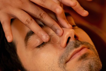 Foto de Hombre caucásico disfrutando de relajante masaje de la cabeza anti-estrés y mimar belleza facial recreación de la piel de ocio en caliente vela iluminación ambiente salón spa en complejo de lujo u hotel. Silencioso. - Imagen libre de derechos