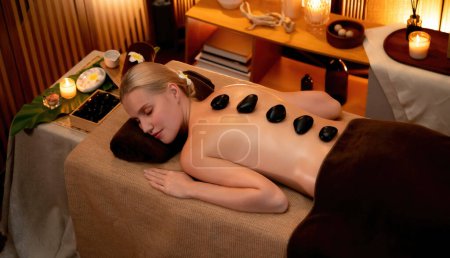 Photo for Hot stone massage at spa salon in luxury resort with warm candle light, blissful woman customer enjoying spa basalt stone massage glide over body with soothing warmth. Quiescent - Royalty Free Image