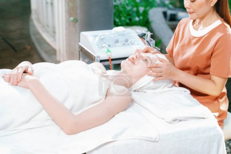 Photo for Portrait of beautiful caucasian woman having facial massage with homemade facial mask while lies on spa bed surrounded by beauty electrical equipment and peaceful nature environment. Tranquility. - Royalty Free Image
