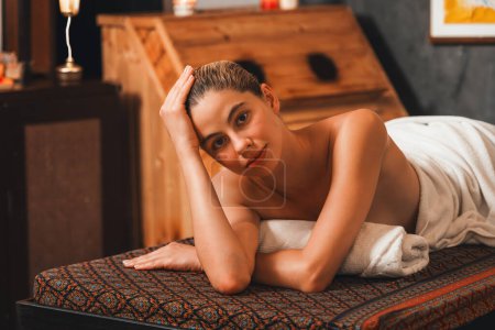 Foto de Mujer joven y bonita se encuentra en la cama de spa frente al gabinete de sauna de madera. Atractivas mujeres caucásicas relajantes en toalla blanca mirando a la cámara mientras esperan un masaje corporal en la sala de sauna.. - Imagen libre de derechos