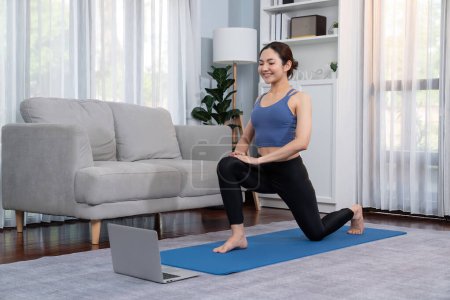 Foto de Mujer asiática en ropa deportiva haciendo ejercicio de yoga en la alfombra de fitness como su rutina de entrenamiento en casa. Cuidado del cuerpo saludable estilo de vida mujer viendo video de yoga en línea en el ordenador portátil. Vigoroso - Imagen libre de derechos
