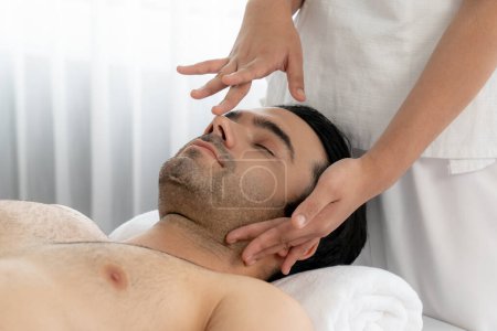 Téléchargez les photos : Homme caucasien appréciant relaxant massage de la tête anti-stress et dorloter beauté faciale loisirs de la peau dans dayspa lumière moderne ambiant au complexe de luxe ou salon de spa de l'hôtel. Tranquillité - en image libre de droit