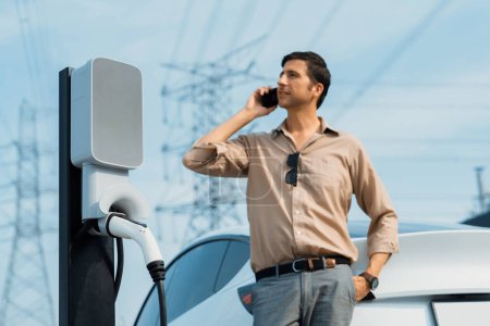 Photo for Man talking on the phone while recharge EV car battery at charging station connected to power grid tower electrical as electrical industry for eco friendly car utilization.Expedient - Royalty Free Image